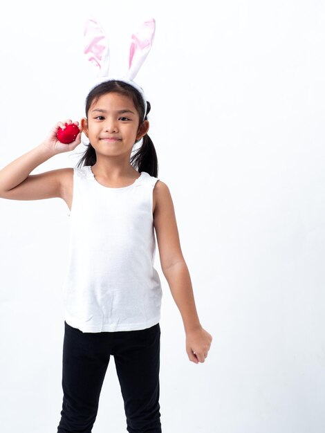 Foto retrato de una niña con una cinta para la cabeza sosteniendo un huevo de pascua mientras está de pie contra un fondo blanco