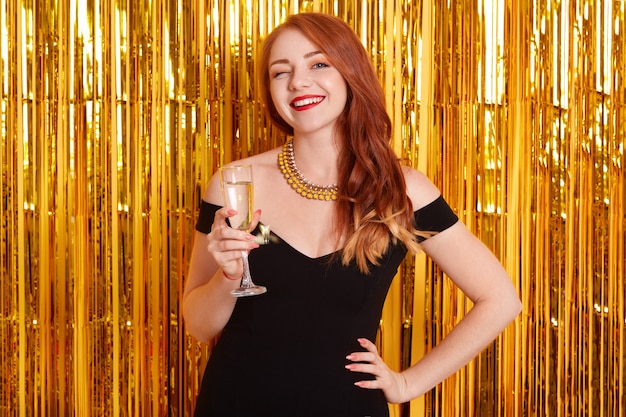 Retrato de niña con copa de champán, posando contra la pared decorada con oropel, atractiva dama con vestido negro, mujer sonriente pelirroja celebrando la fiesta.