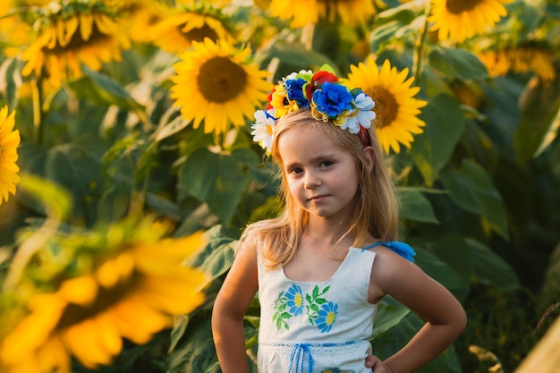 Retrato de la niña con una corona de flores en la cabeza que está de pie con las manos a los lados en el campo de girasoles