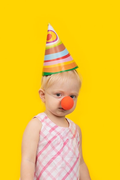 Retrato de niña divertida con gorro de fiesta y nariz de payaso roja en espacio amarillo. Marco vertical.