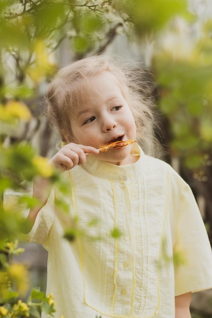 Retrato de una niña divertida con vestido amarillo chupando caramelos en el jardín de flores de fondo