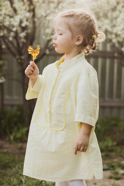 Retrato de una niña divertida con vestido amarillo chupando caramelos en el jardín de flores de fondo