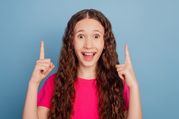 Retrato de niña emocionada indican el espacio vacío de los dedos índices sobre fondo azul.