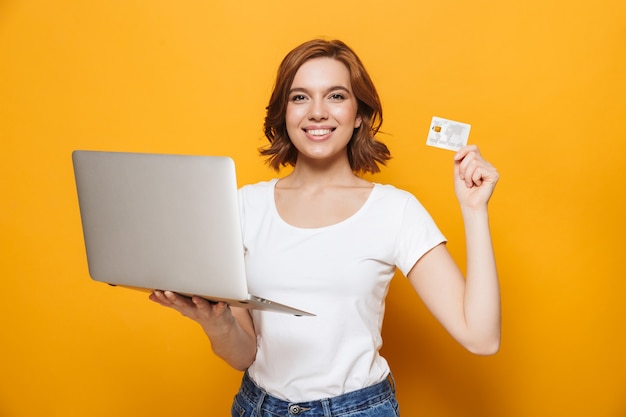 Retrato de una niña encantadora feliz que se encuentran aisladas sobre la pared amarilla, usando una computadora portátil, mostrando la tarjeta de crédito
