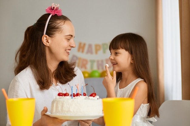 Retrato de una niña encantadora que recibe felicitaciones de una madre encantadora en casa, una madre amorosa que da pastel con velas a una hija feliz y emocionada durante la celebración del cumpleaños