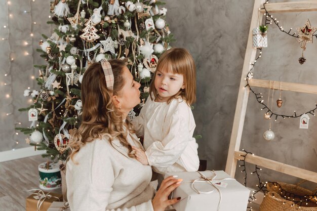 Retrato de una niña encantadora que recibe un regalo de una madre amorosa, sentada cerca del árbol de Navidad. Hermosa mujer hace un regalo de año nuevo a su hijo.