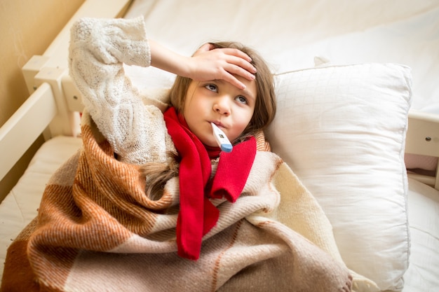 Foto retrato de niña enferma midiendo la temperatura y sosteniendo la mano en la cabeza.