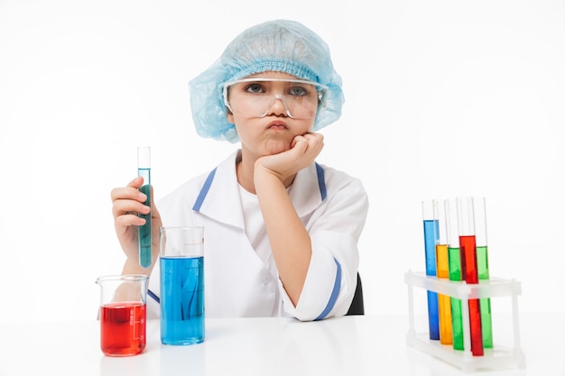 Retrato de niña enfocada en bata blanca de laboratorio haciendo experimentos químicos con líquido multicolor en tubos de ensayo aislados sobre pared blanca