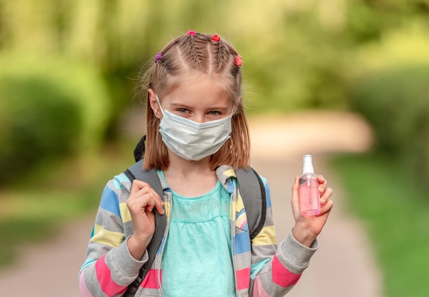 Retrato de niña de la escuela en máscara con desinfectante en la naturaleza