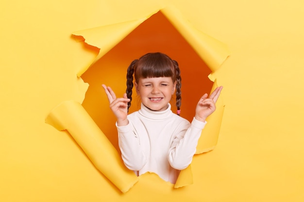 Retrato de una niña esperanzada con trenzas con cuello de tortuga blanco posando en un agujero rasgado de una pared de papel amarillo cruzando los dedos pidiendo esperanzas para que lo mejor mantenga los ojos cerrados