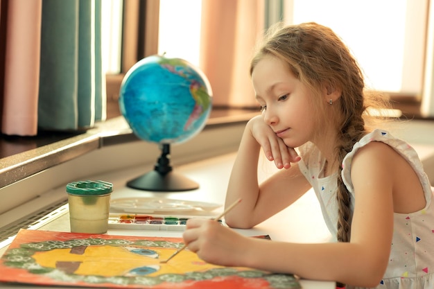 El retrato de una niña está pintando con gouache de colores y una imagen de pincel mientras se sienta en la mesa en el fondo de la habitación