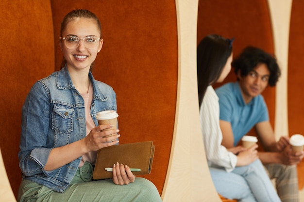 Retrato de niña estudiante inteligente alegre sentado con taza de café y diario en el pasillo de la universidad moderna
