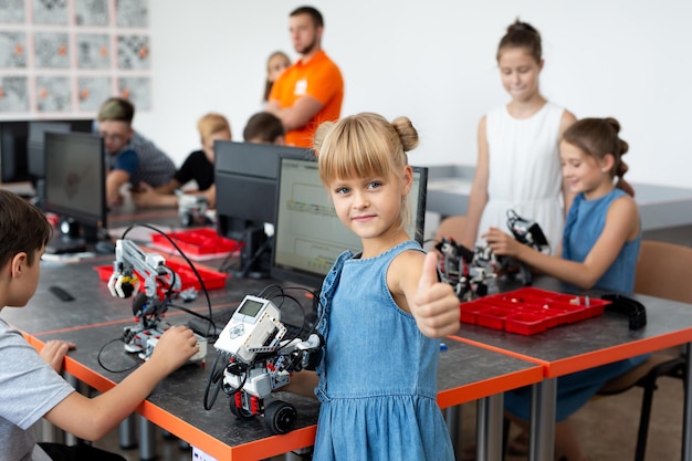 Foto retrato de niña estudiante sostiene un robot en sus brazos, que se ensambló a partir de piezas de plástico programadas en una computadora en una lección de robótica y muestra un pulgar hacia arriba