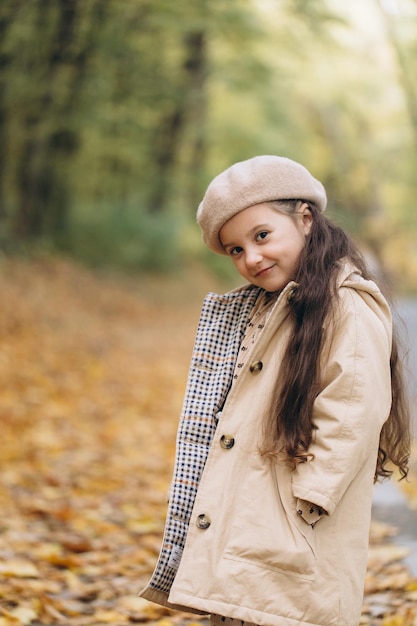 Retrato de una niña feliz con abrigo beige y boina sosteniendo hojas de arce amarillas y pasando tiempo en el parque de otoño