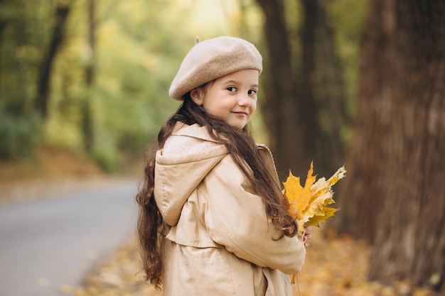 Retrato de una niña feliz con abrigo beige y boina sosteniendo hojas de arce amarillas y pasando tiempo en el parque de otoño