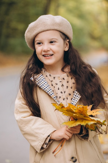 Retrato de una niña feliz con abrigo beige y boina sosteniendo hojas de arce amarillas y pasando tiempo en el parque de otoño