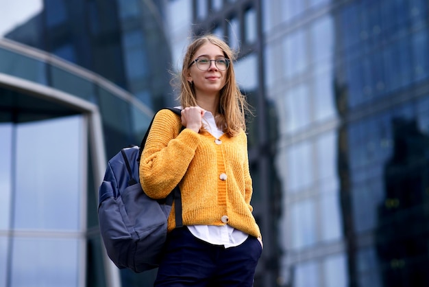 Retrato de niña feliz adolescente hermosa, estudiante universitario o universitario exitoso, joven mujer europea rubia segura de sí misma con mochila en gafas de pie al aire libre en el campus o centro de negocios