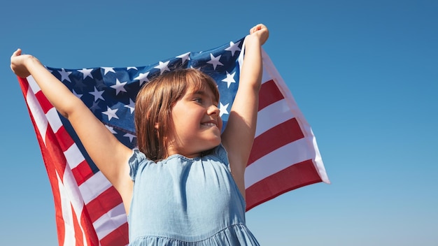 Retrato de una niña feliz con la bandera estadounidense en las manos contra el fondo del cielo
