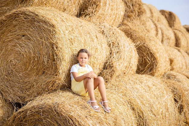 Retrato de una niña feliz y brillante con un trozo de heno en la boca en vestido de verano descansando en un pajar alto y apoyándose en otra pila y otros montones de heno en el fondo Campo lleno de heno dorado