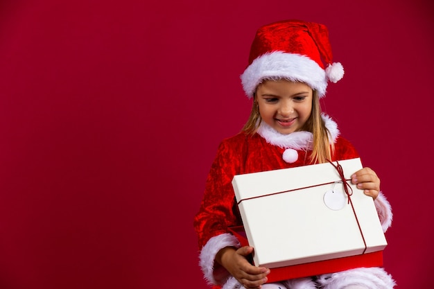 Retrato de una niña feliz con una caja de regalo abierta aislada sobre pared roja, vestida de santa