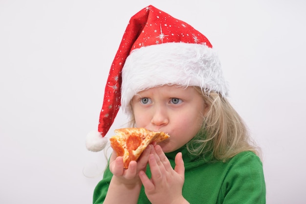 Retrato de niña feliz con gorro de Papá Noel comiendo pizza sobre fondo blanco.