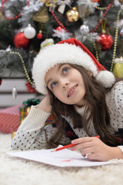 Retrato de niña feliz con gorro de Papá Noel escribiendo carta tirada en el suelo