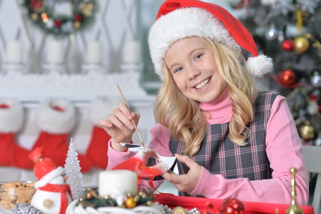 Retrato de niña feliz en Santa hat prapring para Navidad sentado en la mesa y sosteniendo la decoración navideña