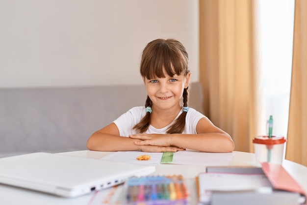 Retrato de niña feliz satisfecha haciendo la tarea escribiendo y leyendo en casa linda niña con cabello oscuro y trenzas mirando a la cámara con expresión positiva aprendizaje a distancia