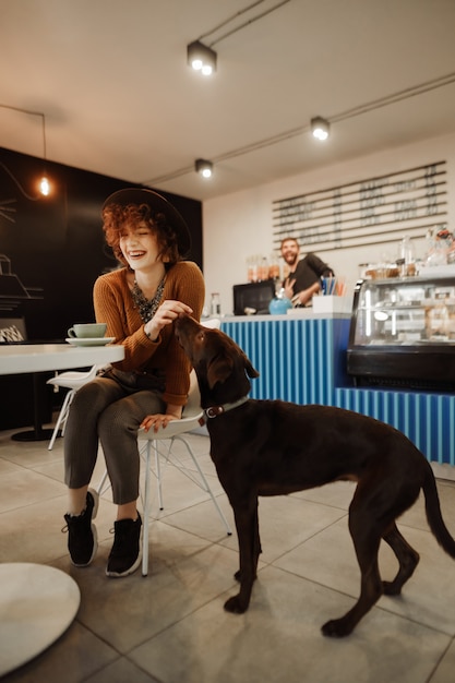 Retrato de niña feliz sentada a la mesa en un acogedor café con una taza de café y jugando con el perro