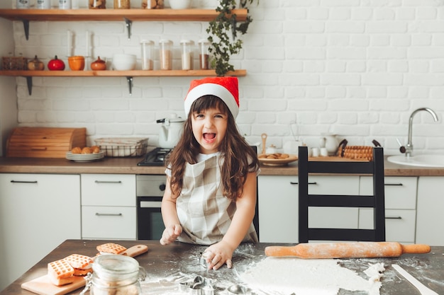 Retrato de una niña feliz con un sombrero de Papá Noel extendiendo masa en la mesa de la cocina un niño preparando galletas de Navidad