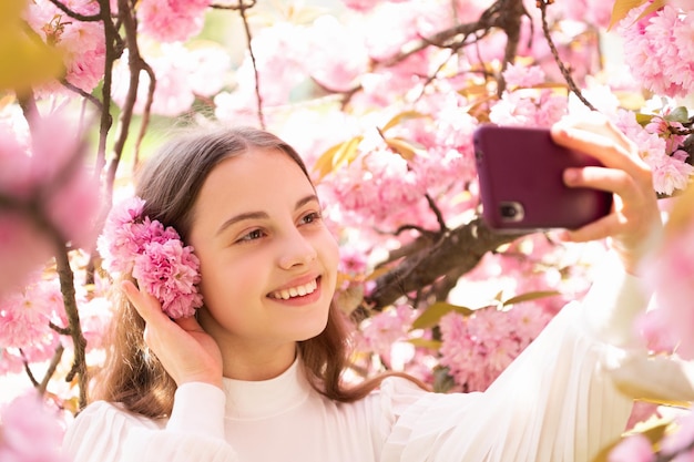 Retrato de niña feliz sonriendo en el teléfono inteligente en el floreciente árbol de sakura en primavera.