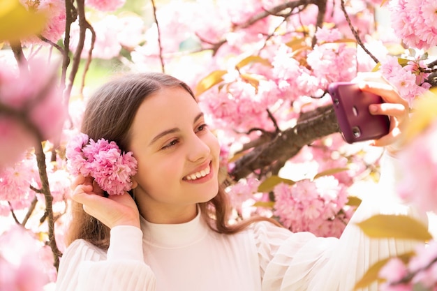 Retrato de niña feliz tomando selfie en árbol floreciente con flores rosas en primavera