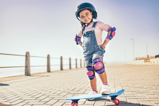 Retrato de niña feliz y de verano con patineta para vacaciones en Los Ángeles con maqueta de cielo Acera de vacaciones y lindo niño negro con casco listo para patinar para el bienestar, la salud y la diversión