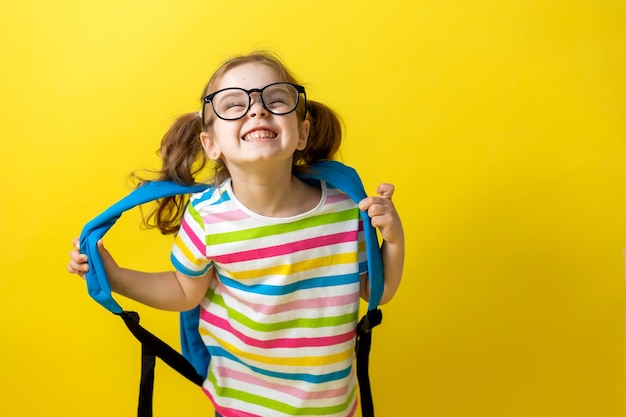 Retrato de una niña con gafas y una camiseta a rayas con una mochila escolar sobre un fondo amarillo. niño alegre tiene prisa por ir a la escuela. concepto de educación. estudio fotográfico, espacio para texto.