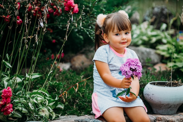 Retrato de una niña hermosa con flores silvestres rosas en la mano sentada cerca de las flores del jardín