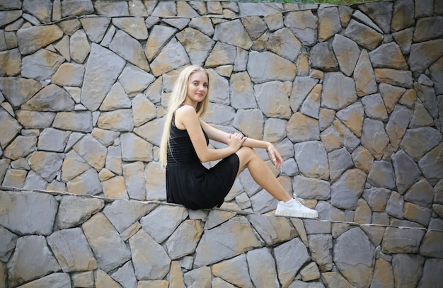 Retrato de niña hermosa rubia con vestido negro contra la pared de piedra como retrato de moda al aire libre.