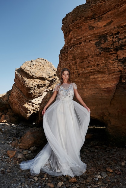 Retrato de una niña en un hermoso vestido de novia posando a un fotógrafo en la playa. La novia está en las rocas