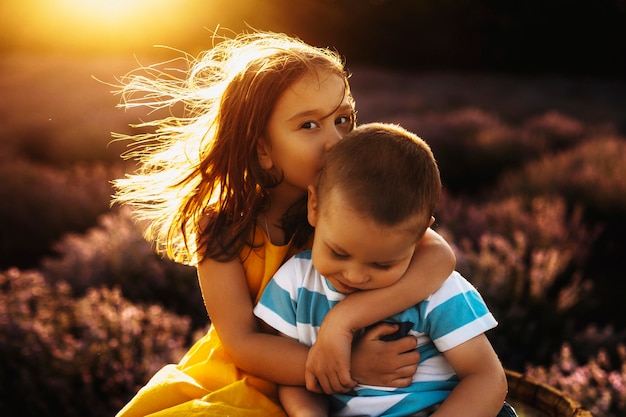 Retrato de una niña increíble vestida con vestido amarillo abrazando y besando a su hermano pequeño en la cabeza mientras mira a la cámara contra la increíble puesta de sol.