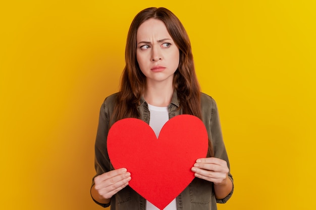 Foto retrato de niña insegura molesta sostenga el símbolo de forma de corazón de papel rojo mirar espacio vacío sobre fondo amarillo