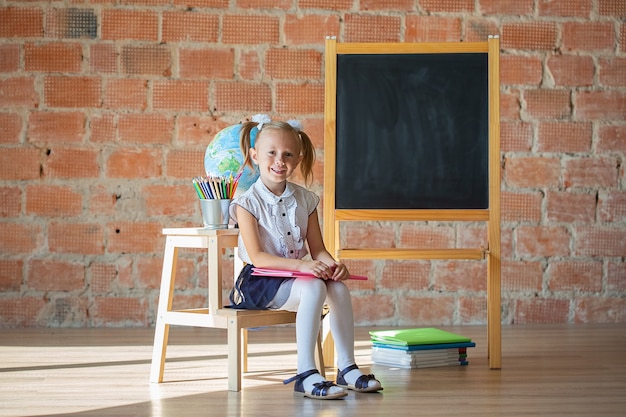 Retrato de niña de jardín de infancia de la escuela caucásica sentado frente a la pizarra con un libro, concepto de regreso a la escuela