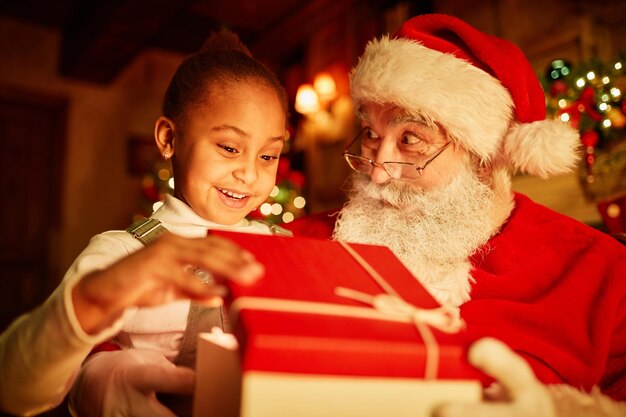 Retrato de niña linda abriendo regalo de Navidad con luz mágica mientras se sienta en el regazo de Papá Noel