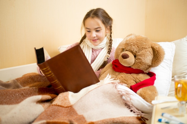 Retrato de niña linda acostada en la cama y libro de lectura para oso de peluche