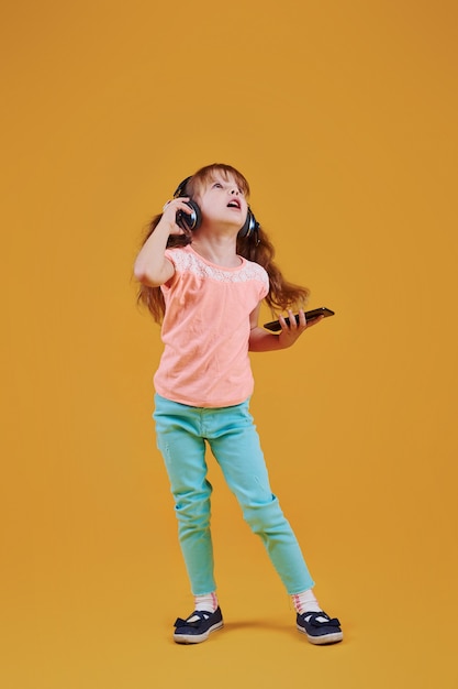 Retrato de niña linda en auriculares y con teléfono en el estudio sobre fondo amarillo.