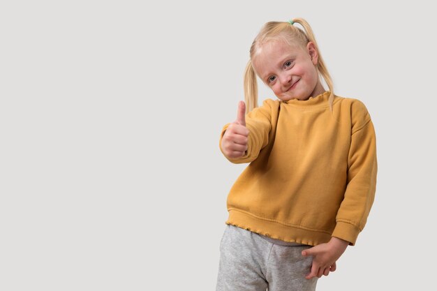 Retrato de una niña linda con cabello rubio en un suéter amarillo que muestra los pulgares en el fondo gris