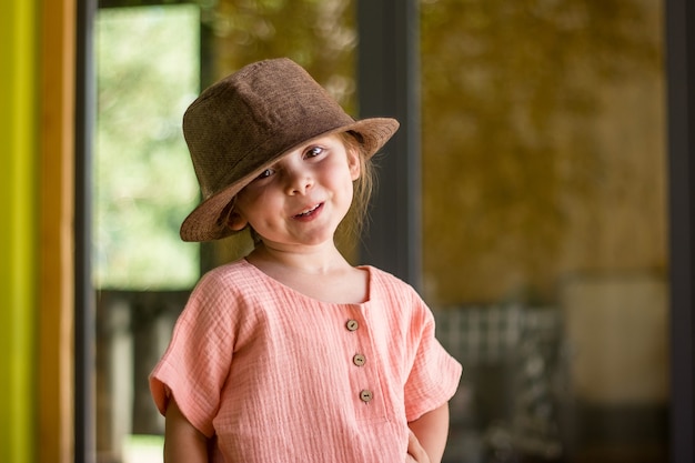 Retrato de niña linda feliz en ropa de muselina y sombrero marrón de estilo rural en el fondo de la ventana