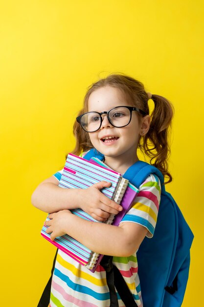 Retrato de una niña linda con gafas en una camiseta a rayas con cuadernos y libros de texto en las manos y una mochila. concepto de educación. estudio fotográfico, fondo amarillo, espacio para texto.