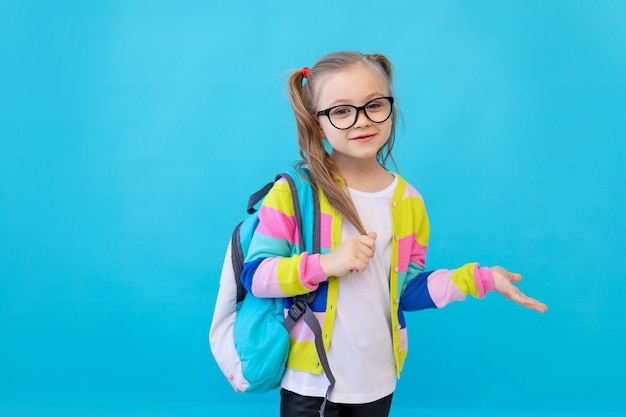 Retrato de una niña linda con gafas en una chaqueta a rayas y una mochila. El concepto de educación. Estudio fotográfico, fondo azul, lugar para texto