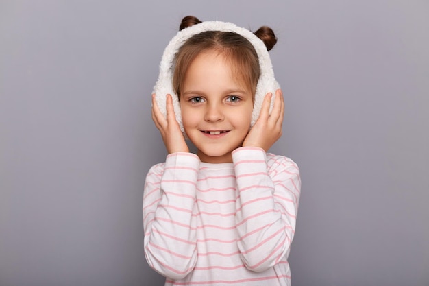Retrato de una niña linda parada en auriculares de piel y mirando a la cámara con una sonrisa expresando emociones positivas usando una camisa a rayas de buen humor