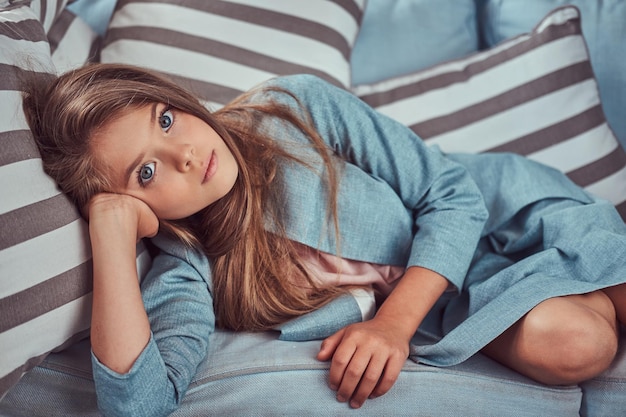 Retrato de una niña linda con el pelo largo y castaño y una mirada penetrante, mirando una cámara, tendida en un sofá solo en casa.