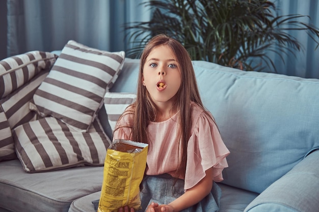 Retrato de una niña linda con el pelo largo y castaño, sentada en un sofá, come patatas fritas.